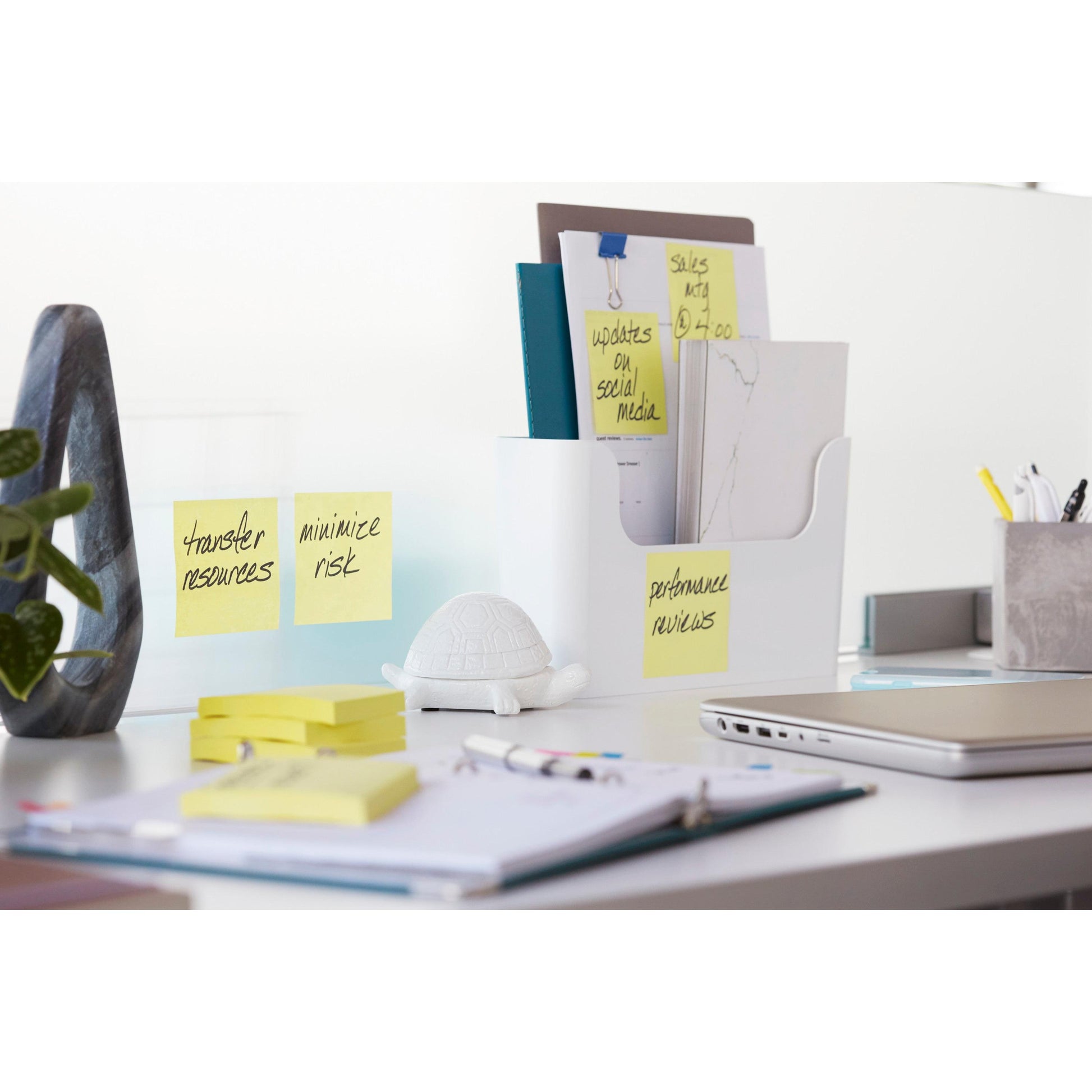 Ein weißer Schreibtisch mit Post-it® Super Sticky Notes (Gelb) der 3M Deutschland GmbH, Dokumenten in einem weißen Organizer, einer Spielzeugschildkröte, einem geschlossenen Laptop, Stiften und einer Pflanze. Auf den Zetteln stehen "Ressourcen übertragen", "Risiko minimieren" und "Leistungsbeurteilung".