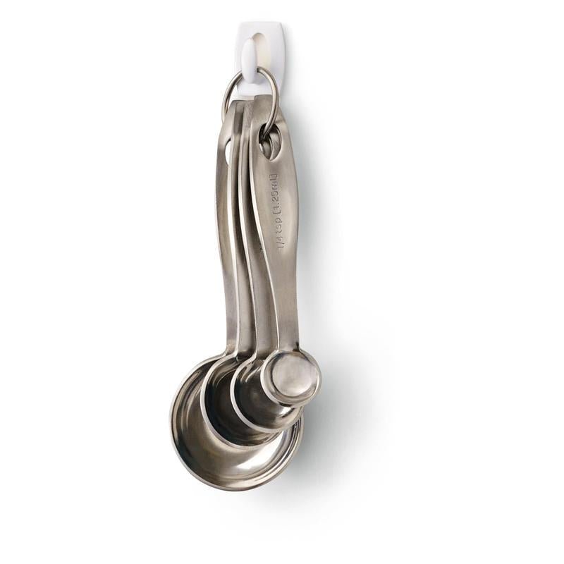 Fyra rostfria mätskedar på en metallring hänger på en 3M Deutschland GmbH Command™ Mini-krok, 17006 - säkert fastsatt utan borrning och lätt att ta bort utan rester från den vita väggen.
