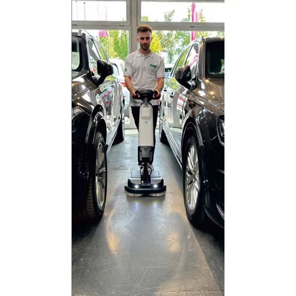 En person i vit skjorta använder Arcora Tiger 40 golvrengöringsmaskinen från Arcora International GmbH mellan två svarta bilar i ett ljust inomhusutrymme med stora fönster och säkerställer en professionell rengöring.