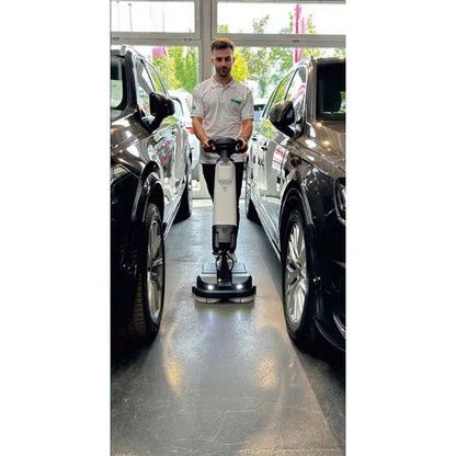 En person i vit skjorta använder Arcora Tiger 40 golvrengöringsmaskinen från Arcora International GmbH mellan två svarta bilar i ett ljust inomhusutrymme med stora fönster och säkerställer en professionell rengöring.
