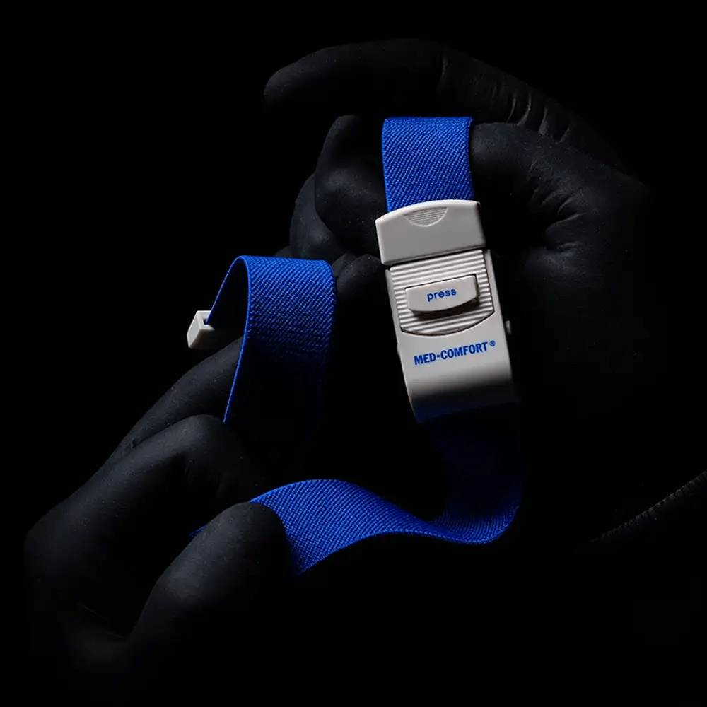En person med svarta handskar håller en blå AMPri MED-COMFORT venstasare, ett oumbärligt instrument för medicinska tillämpningar. På en mörk bakgrund kan ett vitt plastspänne med tryckknapp ses.