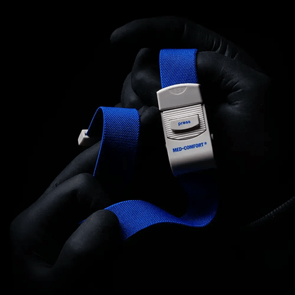 En person med svarta handskar håller en blå AMPri MED-COMFORT venstasare, ett oumbärligt instrument för medicinska tillämpningar. På en mörk bakgrund kan ett vitt plastspänne med tryckknapp ses.