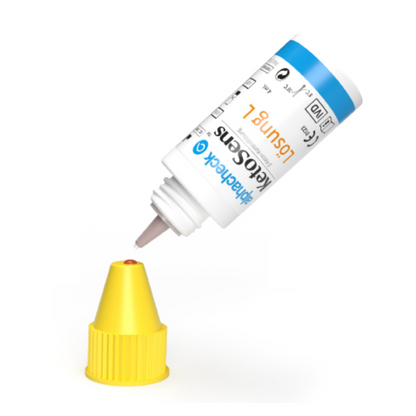 En vit flaska med en blå-gul etikett är upp och ner, där droppmunstycket sitter över ett gult lock. Etiketten innehåller text och symboler som indikerar att det troligen är en produkt Alphacheck KetoSens β-Keton-kontrollösning L+M | Förpackning (2 flaskor) från Berger Med GmbH för användning med Keto-mätinstrument.