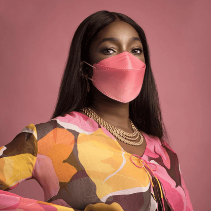 En person bär en partikelfiltrerande Altruan designermask (FFP2-standard) i en elegant rosa nyans och en färgglad topp med abstrakt mönster framför en rosa bakgrund. Hon har långt, mörkt hår och bär flera guldkedjor.