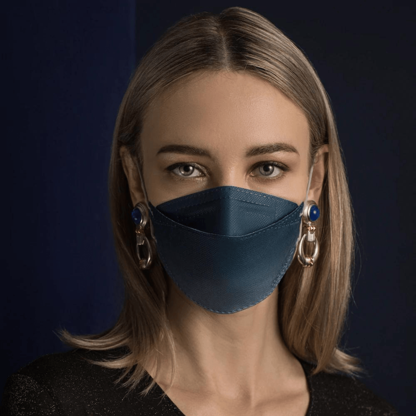 En person med långt hår bär en Altruan designermask partikelfiltrerande i blått, som erbjuder effektivt skydd enligt FFP2-standard. Hon kombinerar den med stora dekorativa örhängen framför en mörk bakgrund och bär en svart skjorta.