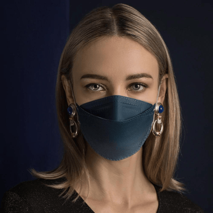 En person med långt hår bär en Altruan designermask partikelfiltrerande i blått, som erbjuder effektivt skydd enligt FFP2-standard. Hon kombinerar den med stora dekorativa örhängen framför en mörk bakgrund och bär en svart skjorta.