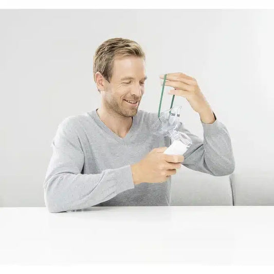 Ein Mann in einem hellgrauen Pullover sitzt an einem weißen Tisch. In einer Hand hält er einen Beurer Inhalator To Go IH 55 | Packung (1 Gerät) und mit der anderen richtet er ein grünes Band. Er lächelt und scheint sich in einem hellen, schlichten Innenraum zu befinden.