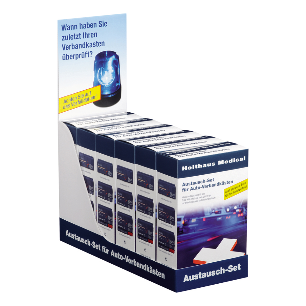 På en utställningsmonter finns flera kartonger med texten „Holthaus utbyteskit för bilförbandslådor DIN 13157 | Förpackning (1 styck)“ från Holthaus Medical GmbH & Co. KG. Montern innehåller en rubrik med en bild av ett blått varningsljus och en tysk text som uppmanar betraktaren att minnas när de senast kontrollerade sina första hjälpen-produkter eller bilförbandslådor.