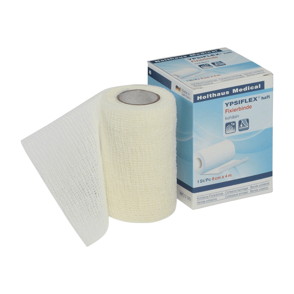 En rulle vit elastisk medicinsk gasbinda, delvis avrullad, ligger bredvid sin förpackningslåda. På lådan syns märket „Holthaus Medical GmbH & Co. KG“ och produktnamnet „Holthaus YPSIFLEX® haft Elastische Mullbinde“, vilket betonar användningen som sårförbandsfixering. Måtten på den självhäftande gasbindan är 8 cm x 4 m.