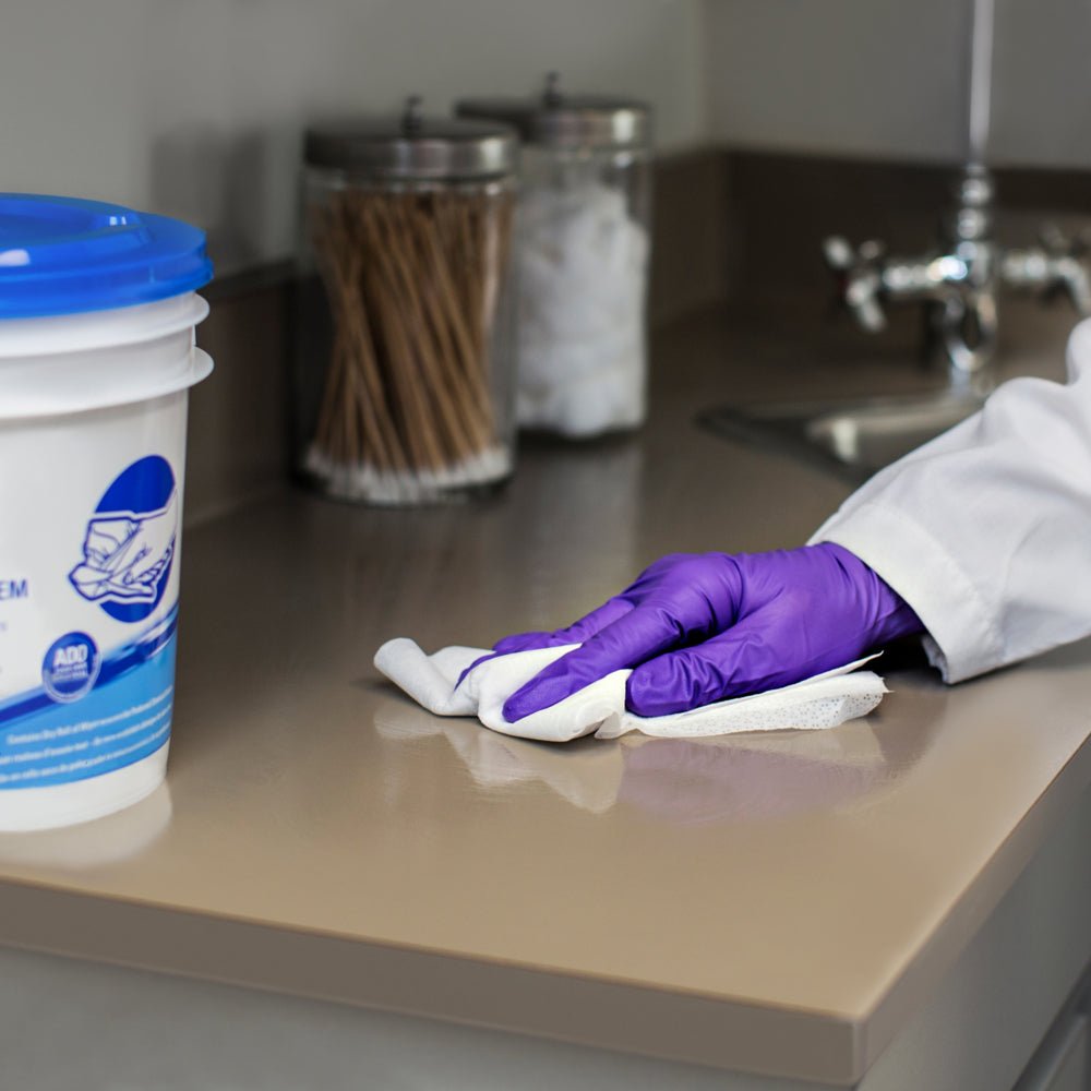 En person med lila handskar torkar av en beige arbetsyta nära diskbänken med WypAll® Wettask™ torkdukar från Kimberly-Clark GmbH (6 rullar x 90 ark), medan burkar med bomullspinnar och -bollar syns i bakgrunden.