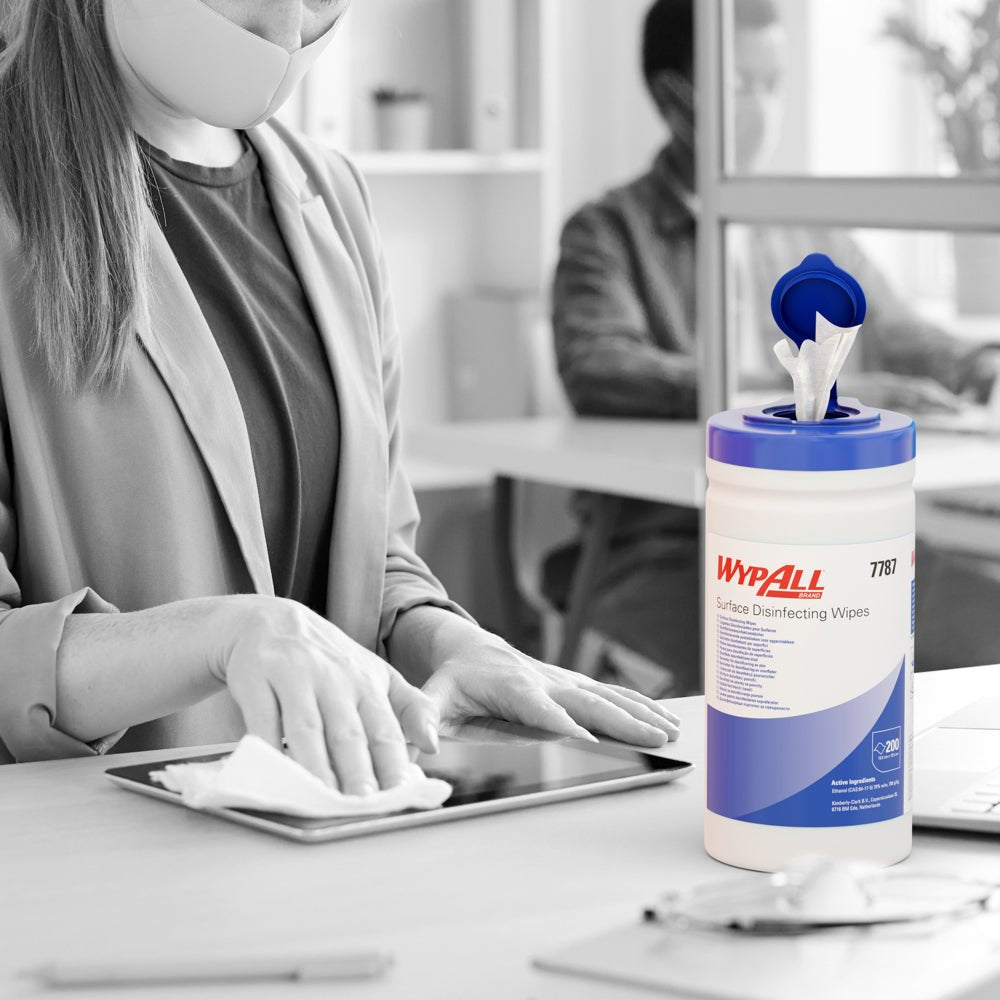 En maskerad person desinficerar med WypAll® ytdesinfektionsservetter (dispenserhink, 200 ark) från Kimberly-Clark GmbH en surfplatta vid ett skrivbord, där hinken är synlig medan en annan person arbetar vid en dator i bakgrunden.