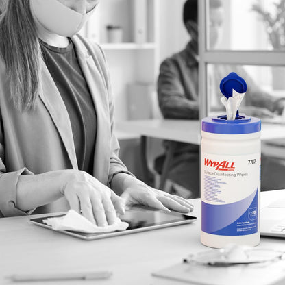 En maskerad person desinficerar med WypAll® ytdesinfektionsservetter (dispenserhink, 200 ark) från Kimberly-Clark GmbH en surfplatta vid ett skrivbord, där hinken är synlig medan en annan person arbetar vid en dator i bakgrunden.