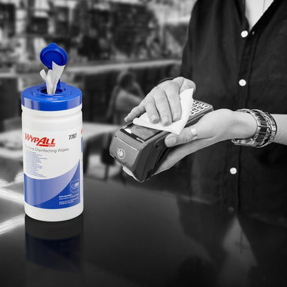 Någon använder en WypAll® ytdesinfektionsservett (Kimberly-Clark GmbH) från en öppen dispenserlåda för att desinficera en kortterminal. Den vita dispenserhinken (200 servetter) står på disken, bakgrunden är suddig.