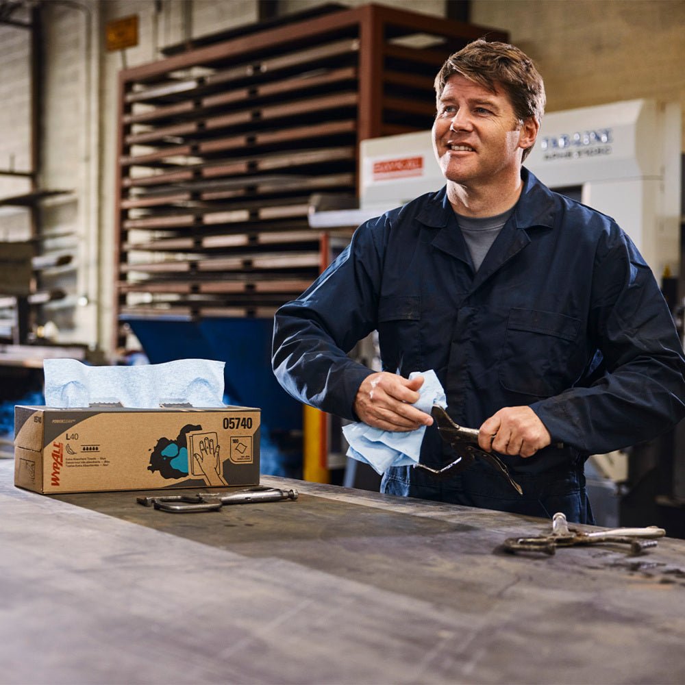En man i en mörkblå overall torkar av ett verktyg i en industriverkstad med en blå engångsduk. Bredvid honom på arbetsbänken står en WypAll® X80 PowerClean BRAG™ Box från Kimberly-Clark GmbH (Vit, 1 Box x 160 ark).
