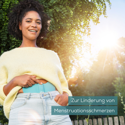 En person i en gul tröja och jeans står utomhus och håller ett Beurer EM 55 Menstrual Relax+ Gel-Pad från Beurer GmbH i handen. I bakgrunden syns grönt genom vilket solljus faller. Texten lyder 'För lindring av menstruationssmärtor'.