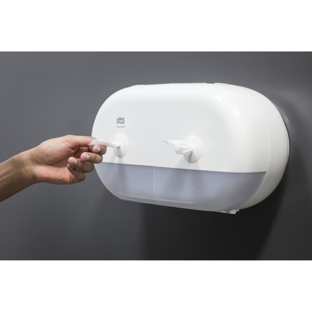 Eine Person zieht ein Papierhandtuch aus einem eleganten weißen Tork SmartOne® 682000 Mini Doppelrollenspender für Toilettenpapier, der über zwei Fächer verfügt und an einer glatten, dunkelgrauen Wand angebracht ist.
