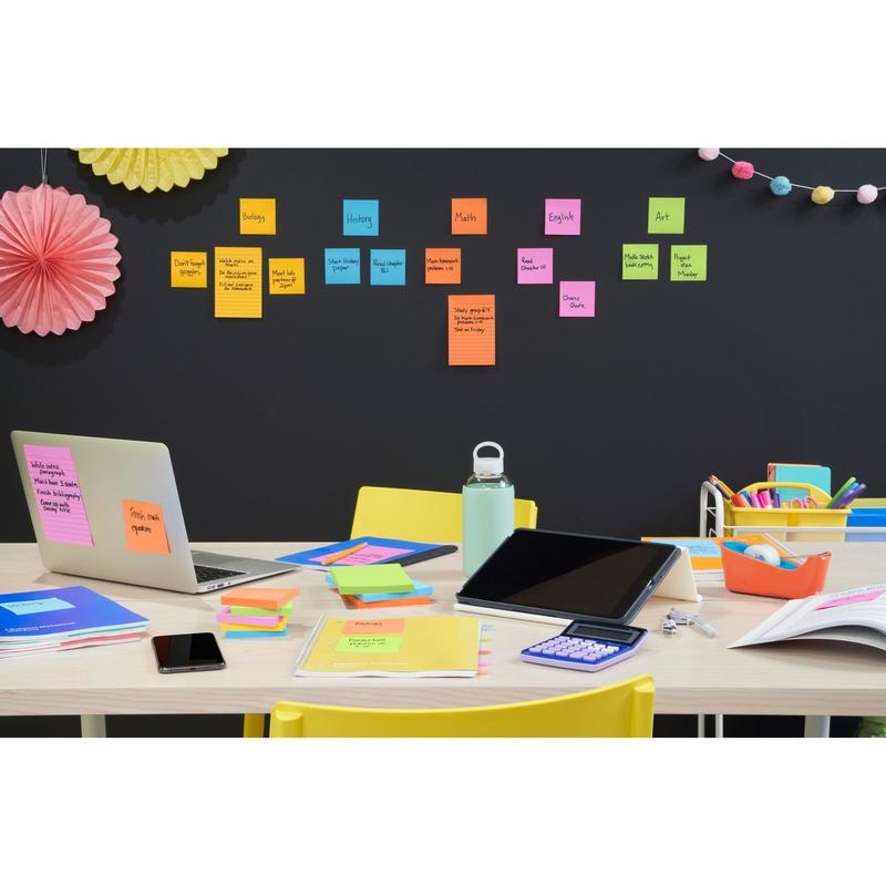 Ett skrivbord med laptop, surfplatta, miniräknare, telefon, vattenflaska och färgglada skrivmaterial. Bakom ordnar Post-it® Super Sticky Notes (76x127mm) från 3M Deutschland GmbH skolämnen på väggen, ovanför hänger dekorationer.
