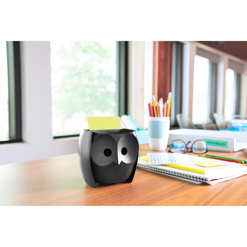 En Post-it® Z-Notes-dispenser, uggla, svart från 3M Deutschland GmbH står på ett träbord med anteckningsböcker, en kopp, färgpennor i en kopp och Post-it® Super Sticky Z-Notes (45 blad/block, 2 block/förpackning); stora fönster ger utsikt över träd.