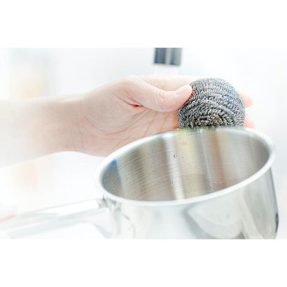 En hand använder en Scotch-Brite® rostfritt stålspiral från 3M Deutschland GmbH, 3 stycken per förpackning, för att avlägsna envisa föroreningar vid rengöring av en rostfri gryta.