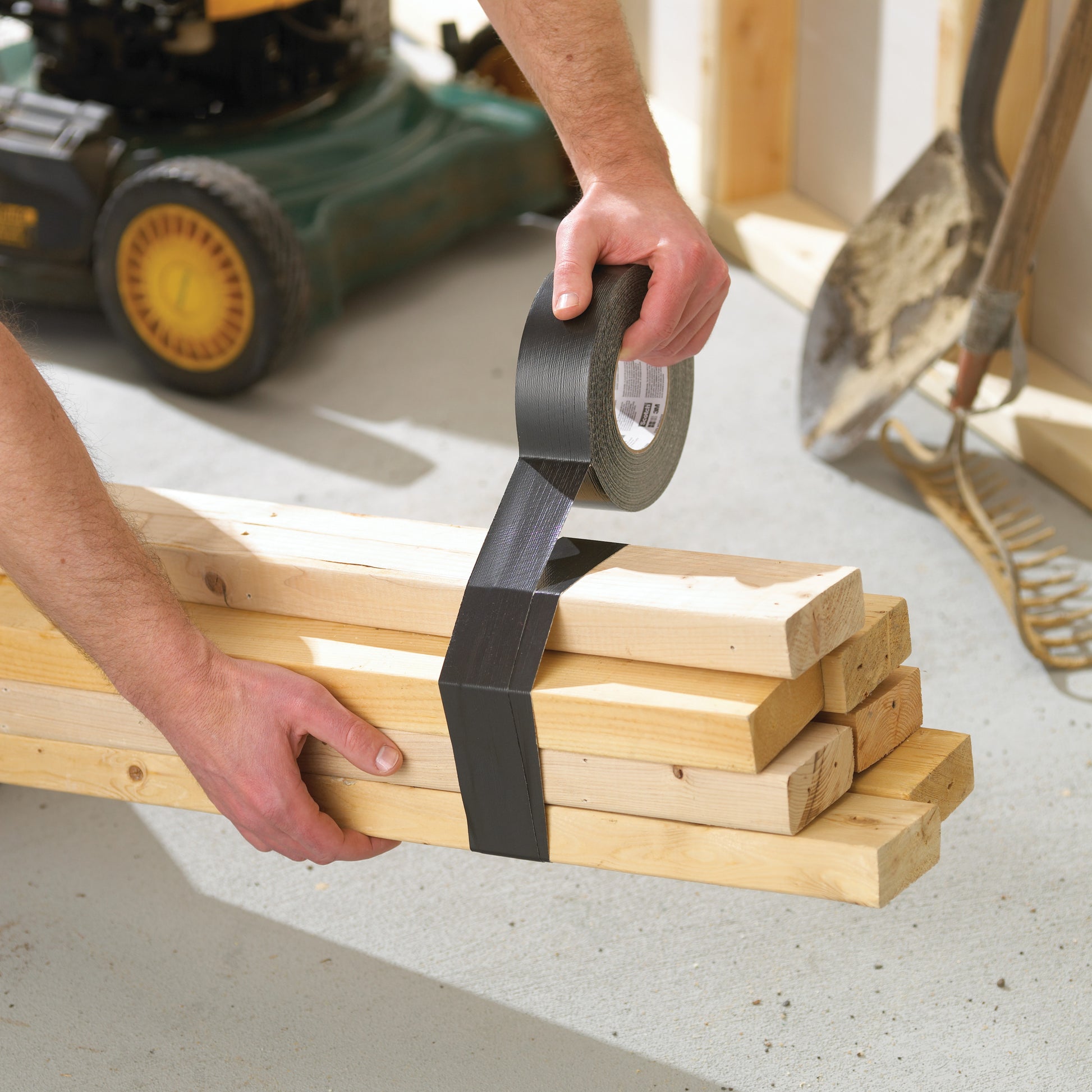 In einer Werkstatt sichert jemand Holzbretter mit Scotch® Extremium™ Ultra Hochleistungs-Klebeband DT17 (10 m x 24 mm) von 3M Deutschland GmbH. In der Nähe stehen eine Schaufel, ein Rechen und ein Rasenmäher.