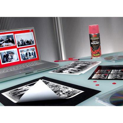 En arbetsyta med en bärbar dator med fotoredigeringsprogram, svartvita utskrifter, en burk 3M™ Scotch® PhotoMount™ spraylim (400 ml, 1 burk, 3M Deutschland GmbH), ett tomt ark för montering, ett genomskinligt CD-fodral och röda klistermärken på ett glasbord.