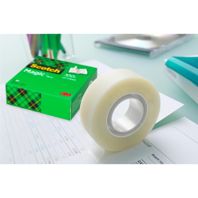En rulle Scotch® Magic™ Osynlig Tejp från 3M Deutschland GmbH i sin gröna förpackning ligger på dokument på ett skrivbord, medan pennor, en anteckningsbok och andra kontorsmaterial är suddiga i bakgrunden.
