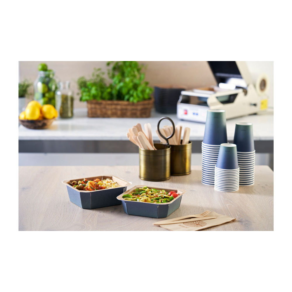 På bordet står två take-away-skålar, flankerade av staplade Duni Urban Kaffeemuggar PAP/PLA från Duni GmbH, träbestick i en hållare och servetter. Citroner, glas och örter i bakgrunden bidrar till den hållbara atmosfären i detta matlagningsområde.